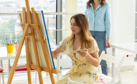 Art school, creativity and leisure concept - student girl or young woman artist with easel, palette and paint brush painting picture at studio.