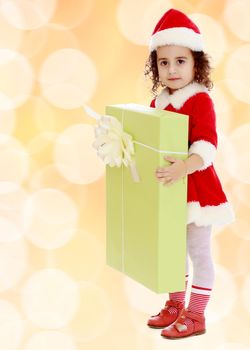 Adorable little curly-haired girl in a coat and hat of Santa Claus,holding a big green box with a gift.Winter brown abstract background with white snowflakes.