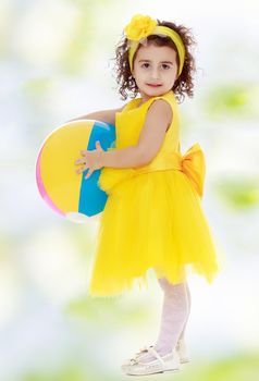 Pretty dark-haired little girl in a bright yellow dress and a bow on my head with a striped ball. Girl posing sideways to the camera in full growth.white-green blurred abstract background