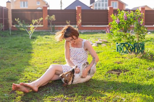 Laughing positive young woman in dress plays with her beloved restless dog sitting in the yard of a country house on a sunny summer day. Family weekend concept