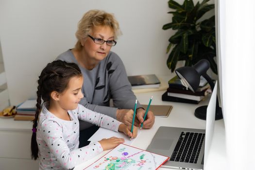 Mature grandmother helping child with homework at home. Satisfied old grandma helping her granddaughter studying in living room. Little girl writing on notebook with senior teacher sitting next to her