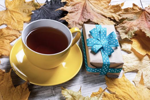 Flat lay autumn composition with gift box and cup of black tea. Time of tea break. Happy thanksgiving congratulation with yellow autumn leaves on vintage wooden desk. Holiday present with blue ribbon