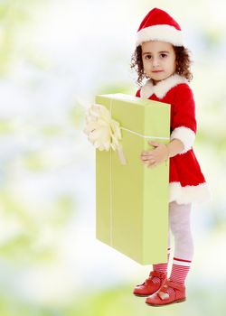 Adorable little curly-haired girl in a coat and hat of Santa Claus,holding a big green box with a gift.white-green blurred abstract background with snowflakes.