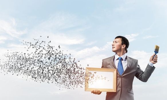 Creative businessman painter holding paint brush and whiteboard in hands. Portrait of happy handsome man in business suit and tie on blue sky background. Various letters flying out from canvas.