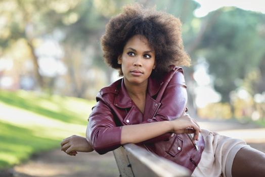 Young black female with afro hairstyle sitting in a bench in an urban park. Mixed woman wearing red leather jacket and white dress with city background.