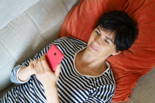 Middle-aged woman checking Social Networks on her smartphone lying on the couch at home.