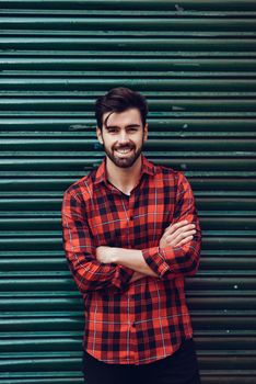 Young bearded man, model of fashion, wearing a plaid shirt with a green blind behind him. Smiling guy with beard and modern hairstyle in urban background.