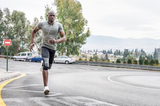 Black man running outdoors in urban road listening to music with white headphones. Young male exercising with city scape at the background.