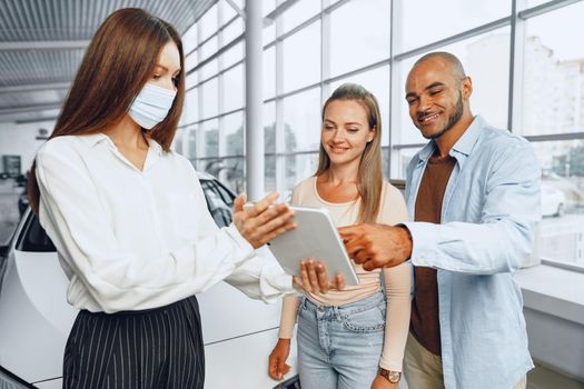 Car saleswoman wearing medical mask shows buyers couple something on digital tablet. .New pandemic job requirements concept