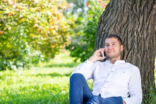 Young man talking on smartphone under tree in park on sunny day. Handsome businessman relaxing outdoors. Smiling man sitting on green grass leaning against tree. Mobile communication concept.