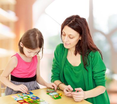 Cute little girl and her mother at the table laid out cards with pictures.In a room with a large semi-circular window.