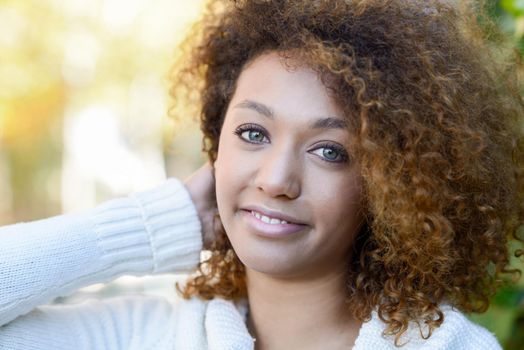 Beautiful young African American woman with afro hairstyle and green eyes wearing white winter dress with autumn leaves in the background.