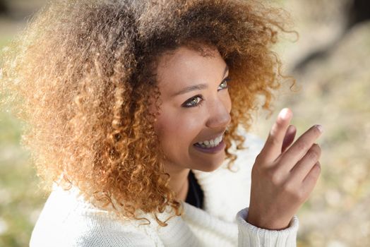 Beautiful young African American woman with afro hairstyle and green eyes wearing white winter dress with autumn leaves in the background.
