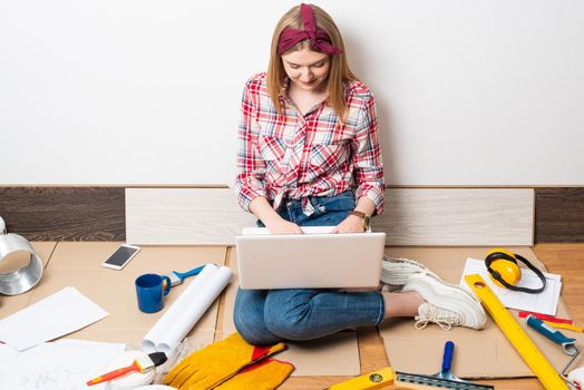 Beautiful girl using laptop at home. Apartment remodeling and house interior renovation concept with copy space. Young attractive woman in red checkered shirt and jeans sitting on floor with computer.