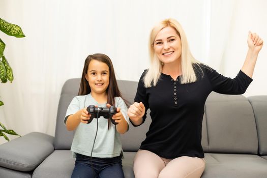 good relationship cute little girl with young mother using joystick playing video game sitting together in living room enjoying family holiday.