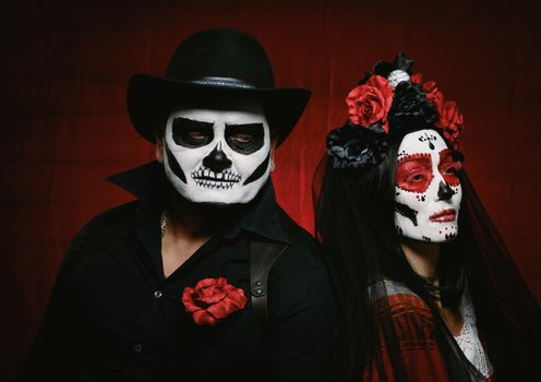 beautiful woman with a sugar skull makeup with a wreath of flowers on her head and a skeleton man in a black hat. Couple on dark red background