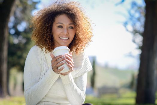 Beautiful young African American woman with afro hairstyle and green eyes wearing white winter dress. Girl drinking coffee in park sitting on grass wearing casual clothes smiling.