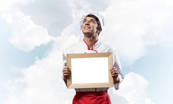 Young male chef holding blank white board for text. Handsome chef in white and red uniform on blue sky background. Cooking classes advertising. Professional catering service concept with copy space.