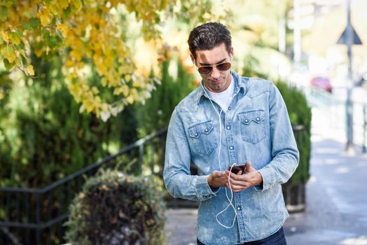 Portrait of young man in urban background listening to music with headphones. Guy wearing casual clothes and aviator sunglasses with a smartphone.