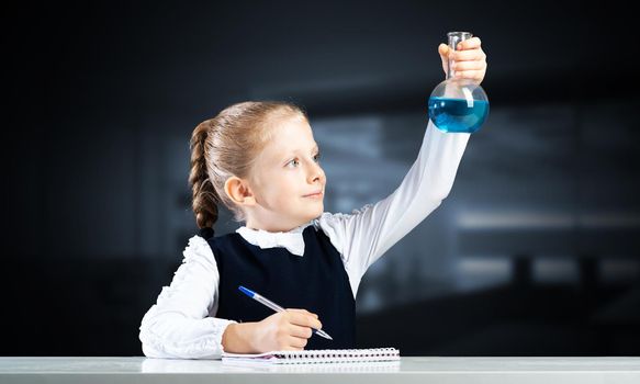 Little girl scientist examining test tube with chemical reagent. Research and education in school. Schoolgirl making experiment in chemistry class. Pupil in schoolwear on deep blue blurred background