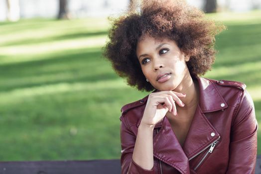 Young black female with afro hairstyle sitting in a bench in an urban park. Mixed woman wearing red leather jacket and white dress with city background.
