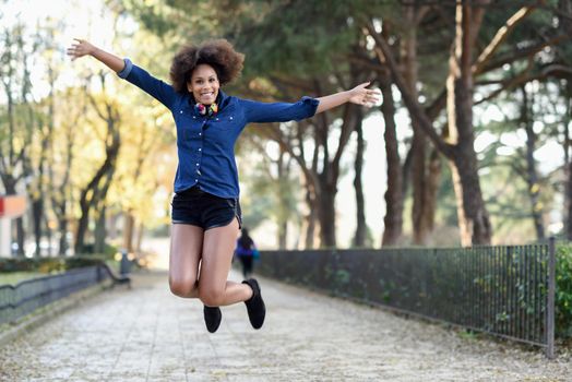 Young black woman with afro hairstyle jumping in urban background with open arms. Mixed woman wearing blue shirt and shorts. Female carrying funny headphones.
