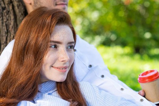 Young couple relaxing with coffee under tree in park on sunny day. Happy couple in love spend time outdoors together. Handsome man and pretty redhead girl sitting on green grass leaning against tree