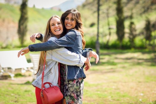 Two happy girls hugging in urban park. Blonde and brunette girls wearing casual clothes outdoors.