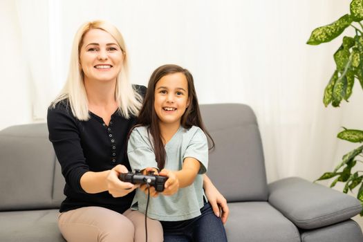 good relationship cute little girl with young mother using joystick playing video game sitting together in living room enjoying family holiday.