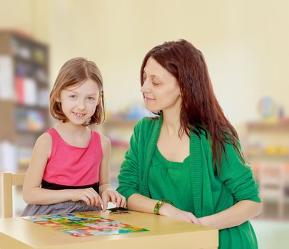 Clever little girl and her teacher at the table laid out cards with pictures.In the children's room where there are shelves with toys.