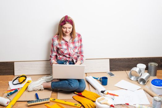 Beautiful girl using laptop at home. Apartment remodeling and house interior renovation concept with copy space. Young attractive woman in red checkered shirt and jeans sitting on floor with computer.