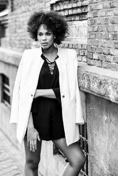 Young black woman with afro hairstyle standing in urban background. Mixed girl wearing white jacket and black dress posing near a brick wall