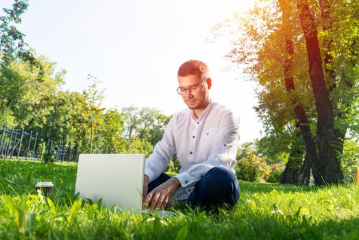 Young businessman using laptop computer on green grass in park. Handsome man in casual wear working with computer outdoors at sunny summer day. Freelance, blogging or remote work concept.