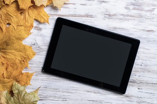 Tablet computer with blank screen lies on vintage wooden desk with bright foliage. Flat lay composition with autumn leaves on white wooden surface. Internet communication and digital technology