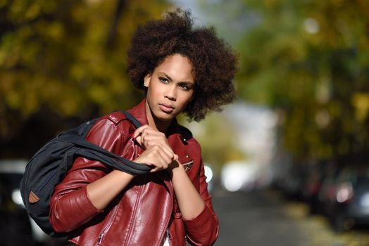 Young black female with afro hairstyle standing in an urban street carrying a bag. Mixed woman wearing red leather jacket and white dress with city background.