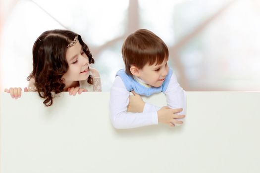 A boy and a girl peeping from behind the white banner.Children look to the side.In a room with a large semi-circular window.