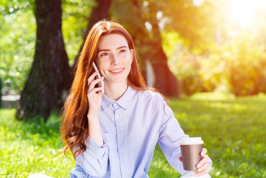 Young redhead woman talking on smartphone in summer park. Beautiful girl relaxing outdoors on sunny day. Charming smiling girl sitting on green grass and drinking coffee. Coffee break concept.