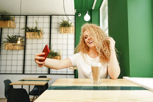 Beautiful curly blonde woman taking selfie while sitting at the table in coffee shop close up