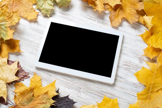 Tablet computer with blank screen lies on vintage wooden desk with bright foliage. Flat lay composition with autumn leaves on white wooden surface. Internet communication and digital technology