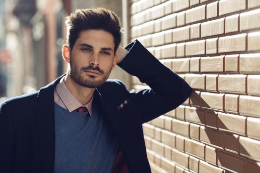 Attractive man in the street wearing british elegant suit. Young bearded businessman with modern hairstyle in urban background.