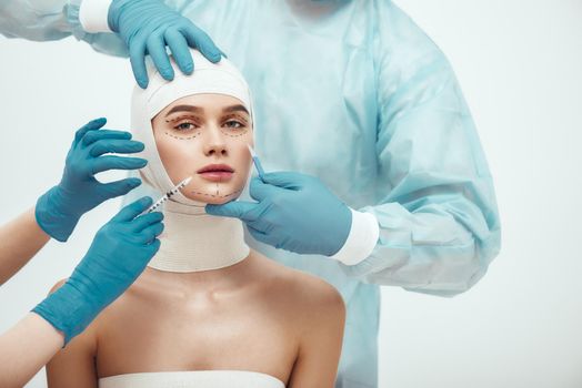 Stop aging. Young attractive woman with head in bandages looking at camera while doctors making plastic surgery operation. Plastic surgeons in blue gloves holding scalpel and syringe near patient face. Facelifting. Cosmetic procedures