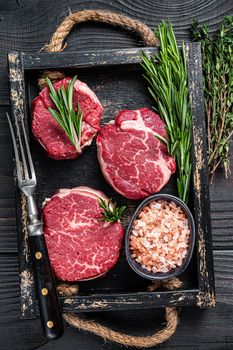 Fillet Mignon tenderloin raw meat veak steaks in wooden tray with herbs. Black wooden background. Top view.