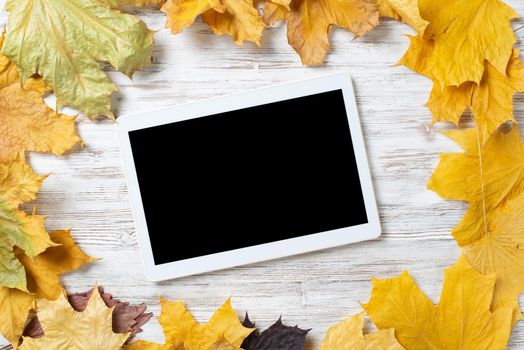 Tablet computer with blank screen lies on vintage wooden desk with bright foliage. Flat lay composition with autumn leaves on white wooden surface. Internet communication and digital technology