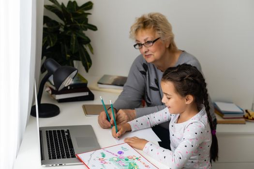 Cute and happy little girl child using laptop computer with her grandma, studying through online e-learning system.