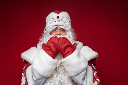 Surprised or shocked Santa in traditional costume and hat looking at camera while covering mouth with both hands. Isolate on red background.
