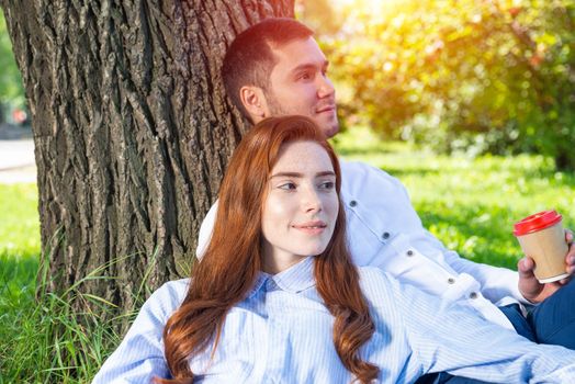Young couple relaxing with coffee under tree in park on sunny day. Happy couple in love spend time outdoors together. Handsome man and pretty redhead girl sitting on green grass leaning against tree
