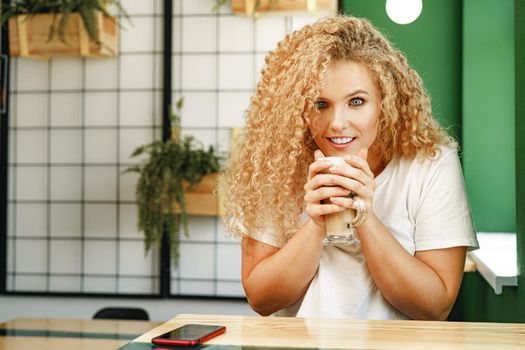 Curly young woman sitting at the table in coffee shop and relaxing with cup of coffee, portrait