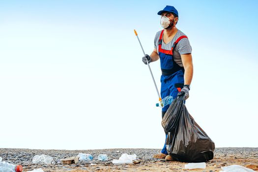 Man volunteer in uniforn collecting garbage on the beach with a reach extender stick