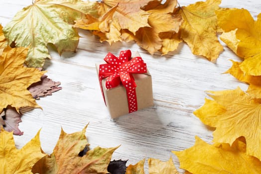 Flat lay cute composition with gift box and yellow autumn leaves. Holiday present decorated red ribbon bow on vintage wooden desk. Happy thanksgiving congratulation. Still life of autumn season.