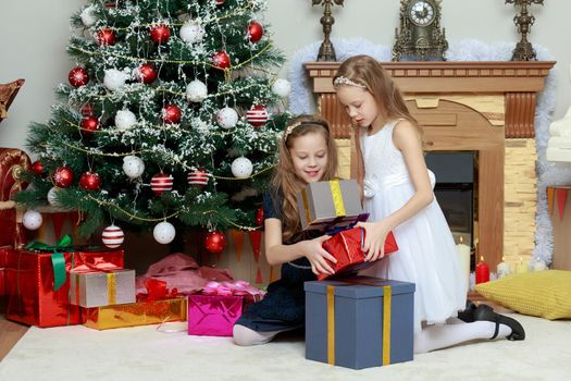 Cute little twin girls, sitting on the floor near the Christmas tree and electric fireplace on which candles are burning. In the hands of the girls boxes with gifts.